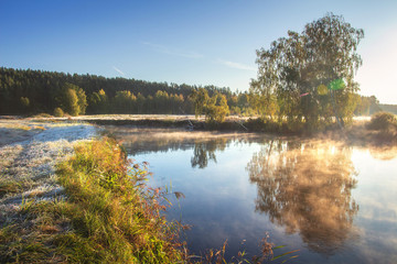 Fototapeta premium Autumn nature. Fall landscape. Calm scene on river side in misty morning at sunrise. Beautiful october foggy morning. Trees on river shore in bright sunlight.