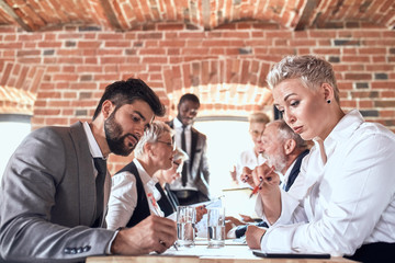 in the foreground caucasian bearded brunet in gray suit man and caucasian blonde woman actively take notes. in background colleagues