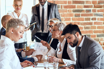 Group of stylishly dressed businessmen sit at table in office and discuss new project. Focused blond caucasian woman who speaks loudly