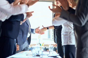 Group of businessmen at table in office. Focuced on shaking hands caucasian man and woman. Others applaud.