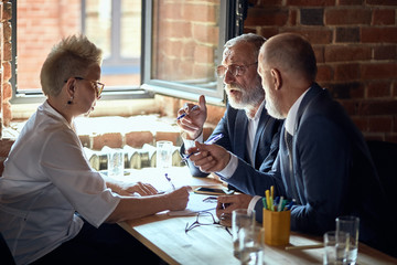 Two adult caucasian blonde men wear blue suits discuss project caucasian blonde woman wear white shirt make notice. On table paper, glasses. Window open.