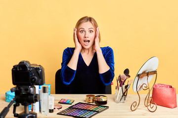 Shocked female model keeps hands near her face, amazed and astonished expression, eyes and mouth wide opened, cant believe in news, sits in front of table with makeup and cosmetics products.