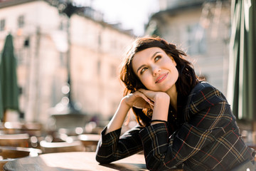 Beautiful woman in a black jacket sits at a table in a city cafe outdoors, smiling and looking up....
