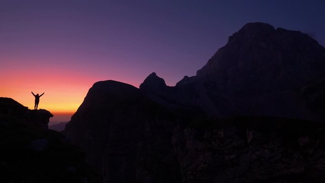 SLOW MOTION, COPY SPACE, SILHOUETTE: Cinematic shot of a cheerful hiker outstretching arms after reaching summit just before sunrise. Joyful trekker celebrates reaching the mountaintop at sunset.