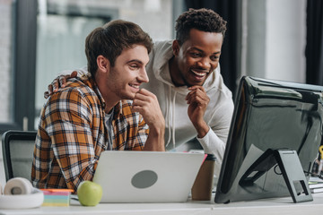 Two cheerful multicultural programmers looking at computer monitor while working in office together