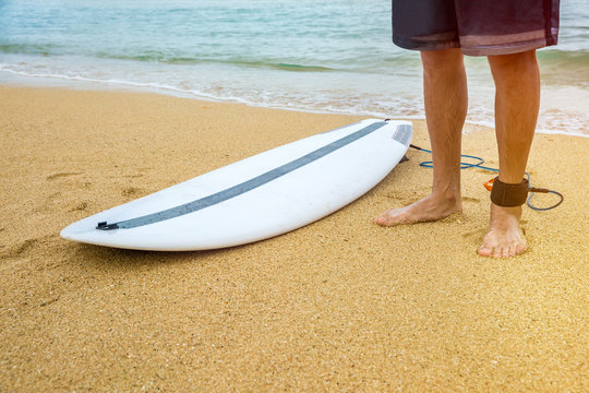 Surfer man stands on sand near the beach with an attached surfboard near the ocean