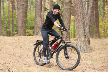 Fototapeta premium Pictiure of biker riding through forest, wearing black cycling clothes, cap and sneakers, having happy facial expression, sporty male spending leisure time in open air, riding around trrees.