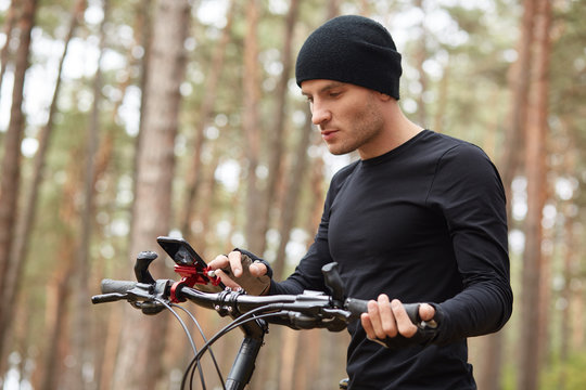 Outdoor picture og attractive young guy holding mobile phone in hands, male traveling on bike through forest, finding right way via smartphone, internrt or navigator, spending free time in open air.