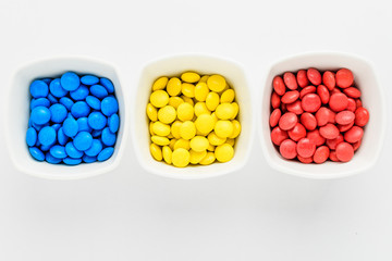 Three squared bowls with small red, yellow and blue coated chocolate candies similar to m&ms in a...