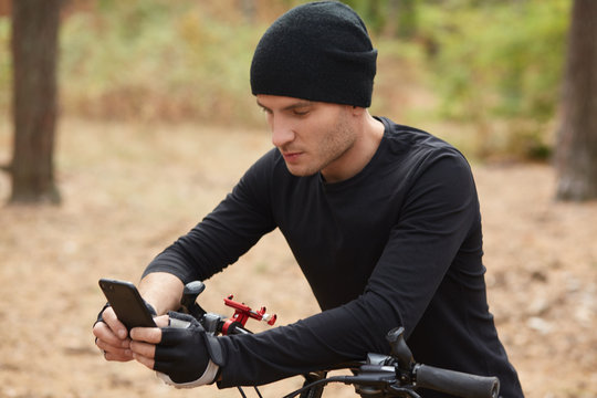 Outdoor Shot Of Bikepacker Stops In Middle Of Forest And Using His Phone For Finding Right Way, Checking Social Networks Or Messaging His Friends, Guy Wears Black Sportwear And Cap. Lifestyle Concept.