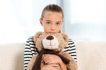 cute kid holding teddy bear while looking at camera in living room