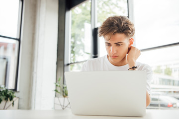 pensive teen boy in white t-shirt using laptop at home