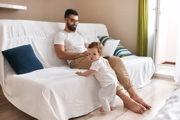 Fototapeta premium smiling young father enjoying working on laptop, while his baby leaning on the sofa and looking at the camera. close up photo