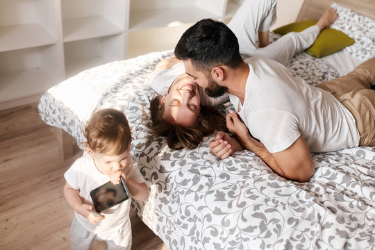 young couple is kissing while lying on the bed, their little kid holding smart phone, using it. close up photo
