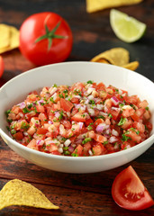 Mexican Tomato Salsa in white bowl with lime, red onion, jalapeno pepper, parsley and tortilla chips on wooden table