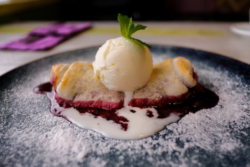 Cherry strudel cake served with ice cream on wooden table at restaurant. Dessert sprinkled with sugar on a blue plate.