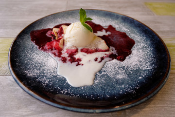 Cherry strudel cake served with ice cream on wooden table at restaurant. Dessert sprinkled with sugar on a blue plate.