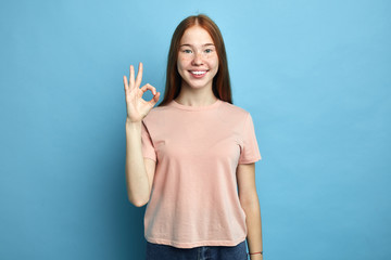 positive female student with freckles showing ok sign with fingers. close up portrait, isolate blue background, studio shot, agreemnet. body language