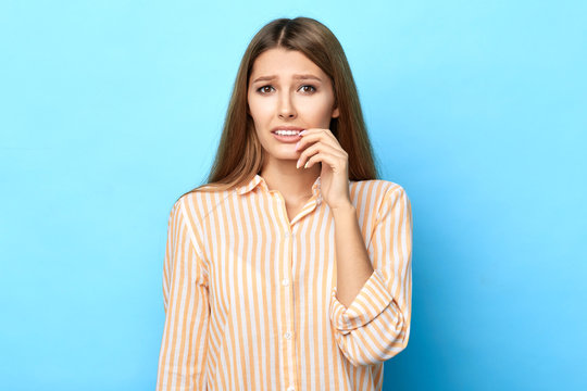 Close Up Portrait Of A Pensive Worried Scared Young Woman Biting Her Nails Isolated Over Blue Background, Studio Shot. I Am Sorry