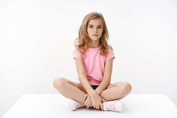 Serious indifferent cute blond little girl sitting on floor, legs crossed, look bored and unamused,...