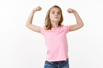 Cute charismatic lovely blond girl want grow up strong and empowered, showing strength raise hands brag muscles and biceps, pull serious confident expression, look camera ambitious, white background