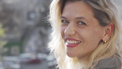 Lovely mature woman smiling to the camera over her shoulder. Happy blond haired woman observing busy city streets, smiling to the camera joyfully. Urban lifestyle concept