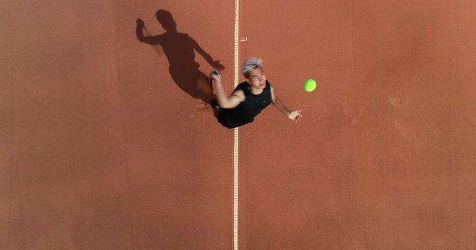 A platinum haired mixed race tennis player serves filmed from above.