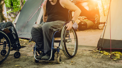 Disabled man resting in a campsite with friends. Wheelchair in the forest on the background of tents