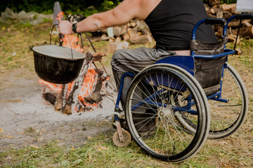 Fototapeta na wymiar Disabled man resting in a campsite with friends. Wheelchair in the forest on the background of bonfire. Barbeque. Camping.