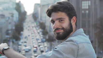 Handsome bearded man smiling to the camera over his shoulder. Young male hipster looking away at busy city streets, smiling joyfully to the camera. Success, achievement concept