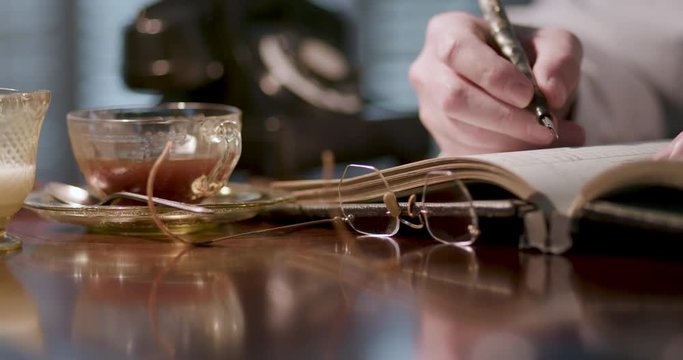 A man writing in a ledger with a classic ink fountain pen with vintage eyeglasses and depression period glass coffee cup near by.