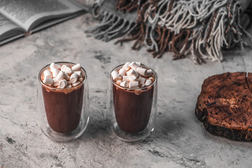 Two glasses of hot cocoa and marshmallow, a wooden stump-stand for cups, a book and a blanket