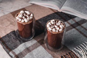 Two glasses of hot cocoa with marshmallows on a brown wool blanket next to an open book