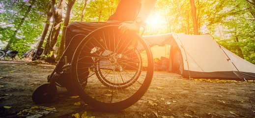 Disabled man resting in a campsite with friends. Wheelchair in the forest on the background of tents
