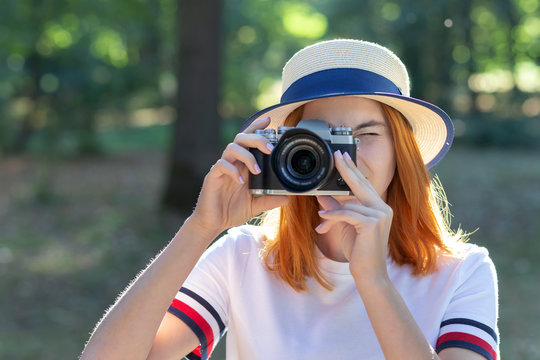 Pretty teenage girl with red hair taking picture with photo camera in summer park.
