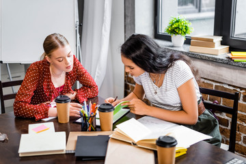 attractive multicultural friends sitting at table and studying in apartment