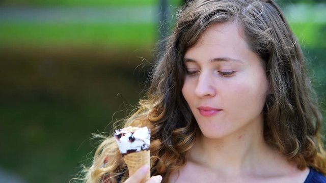 Woman eating one vanilla chocolate peanuts ice cream gelato cone with bokeh background of park in Europe city during sunny summer day