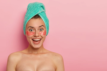 Studio shot of positive European woman wears collagen patches under eyes, has beauty treatments, stands naked indoor, has broad smile, appealing look, isolated over pink background, copy space