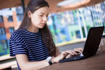 Close-up, portrait of a serious, gently smiling girl student, freelancer, blogger working at a laptop at a wooden table.