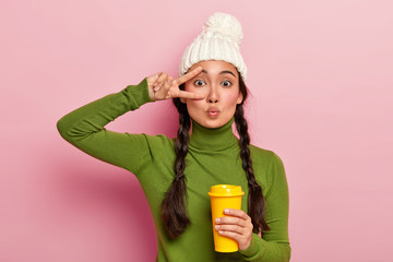Image of young Asian girl keeps lips folded, makes peace gesture, wears white warm winter hat,...