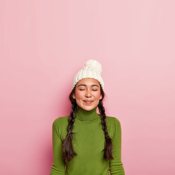 Vertical image of pleased dark haired girl keeps eyes closed, thinks about pleasant meeting with friend, wears white hat and green turtleneck, stands against pink background, free space above