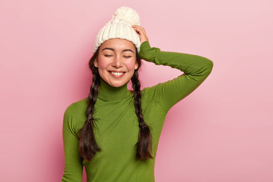 Portrait of beautiful hipster girl, keeps eyes shut, dressed in green poloneck and hat, satisfied after spending free time with close person remembers pleasant moment during romantic date. Cold season