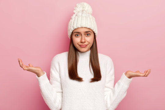 Image of hesitant European girl shrugs shoulders, being questioned what to do, looks helpless, doesnt know how to act in difficult situation, wears white hat and knitted sweater, looks confusingly