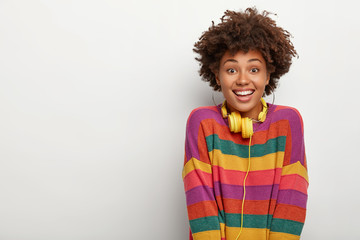 Portrait of pleased millennial Afro girl enjoys spare time, listens music via headphones, looks...