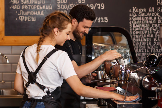 Two young smiling barista at work. Professional barista team brewing coffee using coffee machine in coffee shop. Happy young man and woman developing own coffee business. Coffee shop concept.