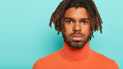Fototapeta premium Headshot of serious youngster has dark eyes, healthy groomed skin, has dreadlocks, thick bristle, has self assured gaze at camera, wears comfortable clothes, isolated on blue wall, free space for text