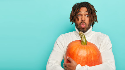 Photo of astonished black man holds big orange pumpkin, scared by something, has specific hairstyle, surprised with rich seasonal harvest, wears white sweater. People and Halloween preparation concept