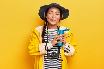 People and tourism concept. Satisfied attractive female with pigtail, drinks hot coffee or tea from flask, wears casual waterproof clothes, retro camera hanging on neck, likes adventures and traveling