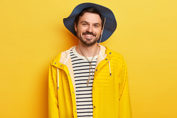 Horizontal shot of pleased Caucasian man wears hat, yellow raincoat, spends leisure time actively, poses indoor, expresses good emotions, has stubble, stands indoor. Happy male model travels alone