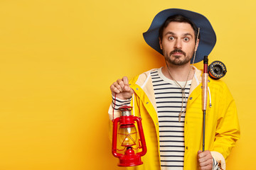 Surprised fisherman holds fishing rod and kerosene lamp, has overnight fishing trip, wears hat and raincoat, poses over yellow background, copy space for your advertising area. Angling concept